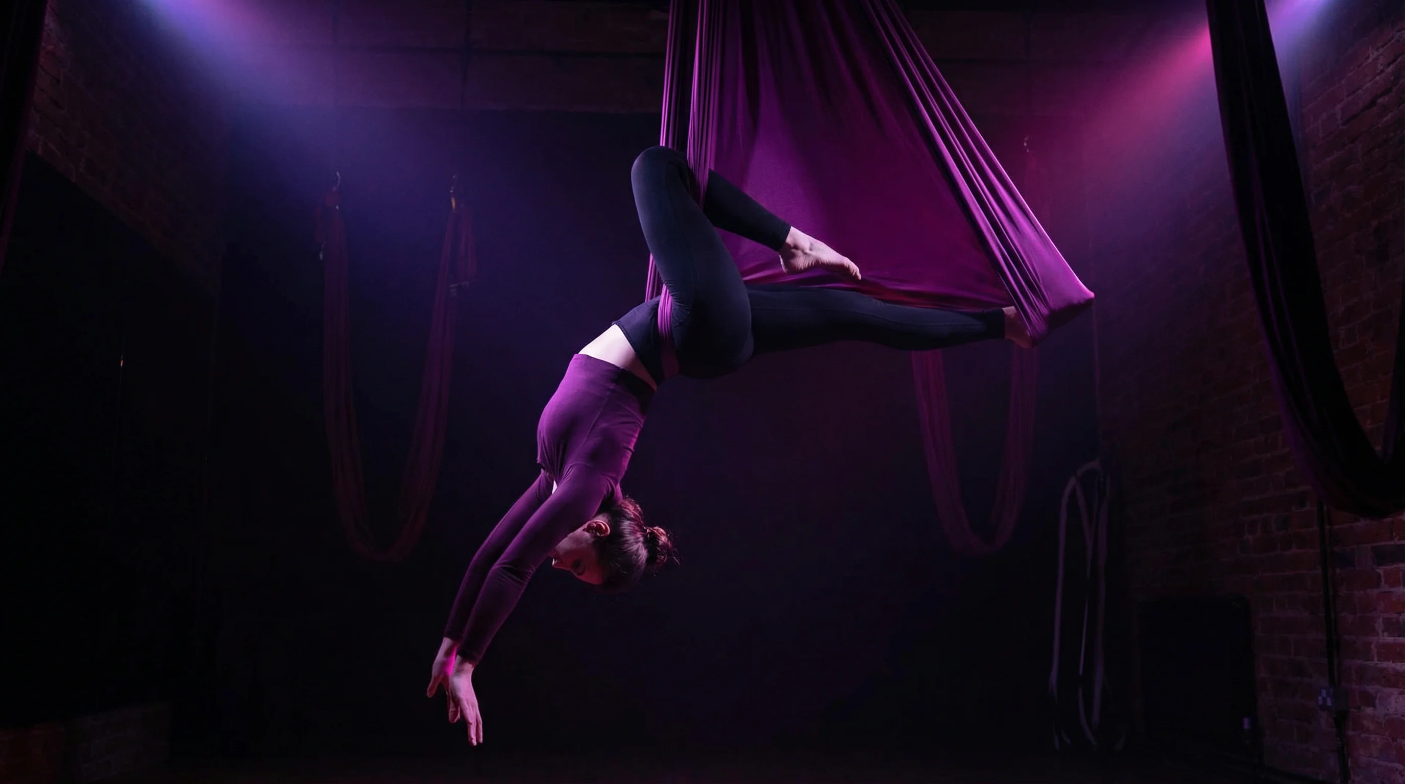 A woman suspended in a deep purple silk aerial yoga hammock performing an inverted pose in a dark studio