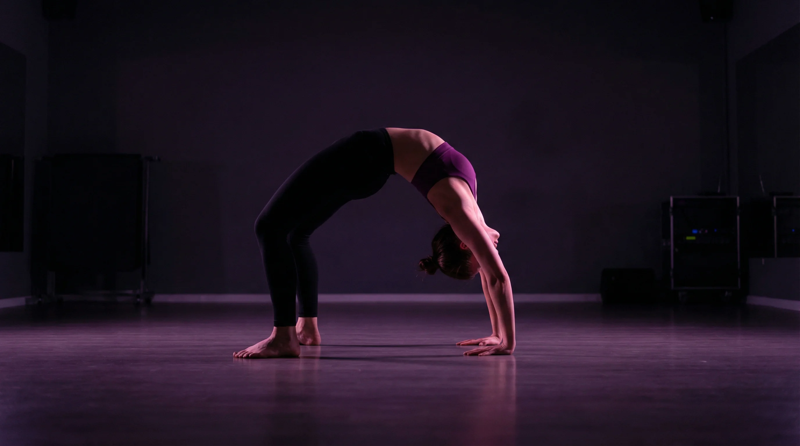 A female dancer performing a full wheel pose backbend on a dark studio floor with purple and magenta lighting