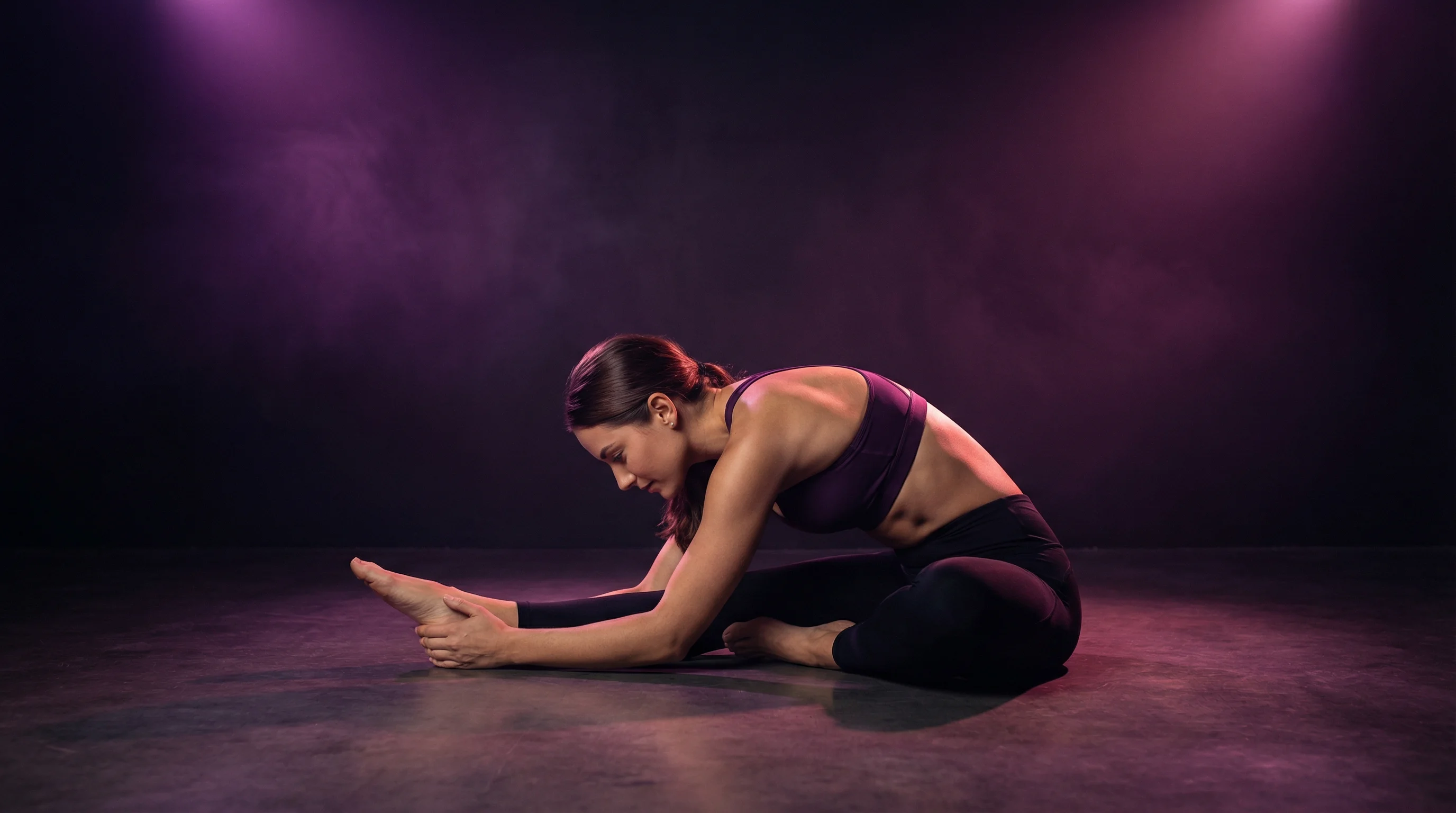 A woman performing a seated forward fold stretch on a dark studio floor with dramatic purple and magenta lighting