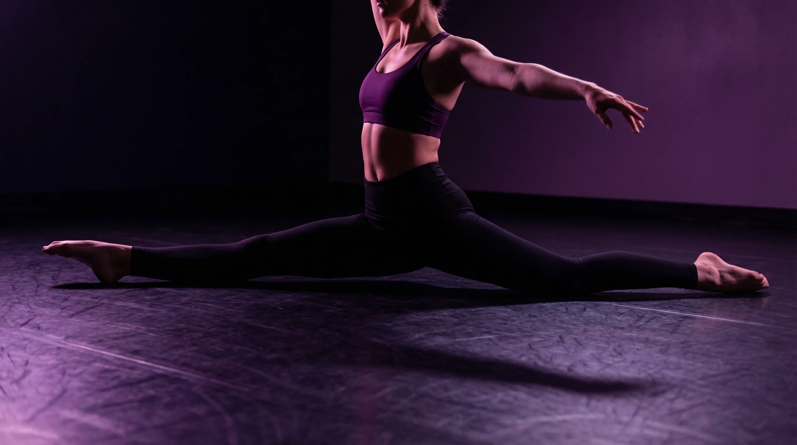 Female dancer in a perfect front splits position on a dark studio floor, right leg extended forward and left leg extended back at 180 degrees