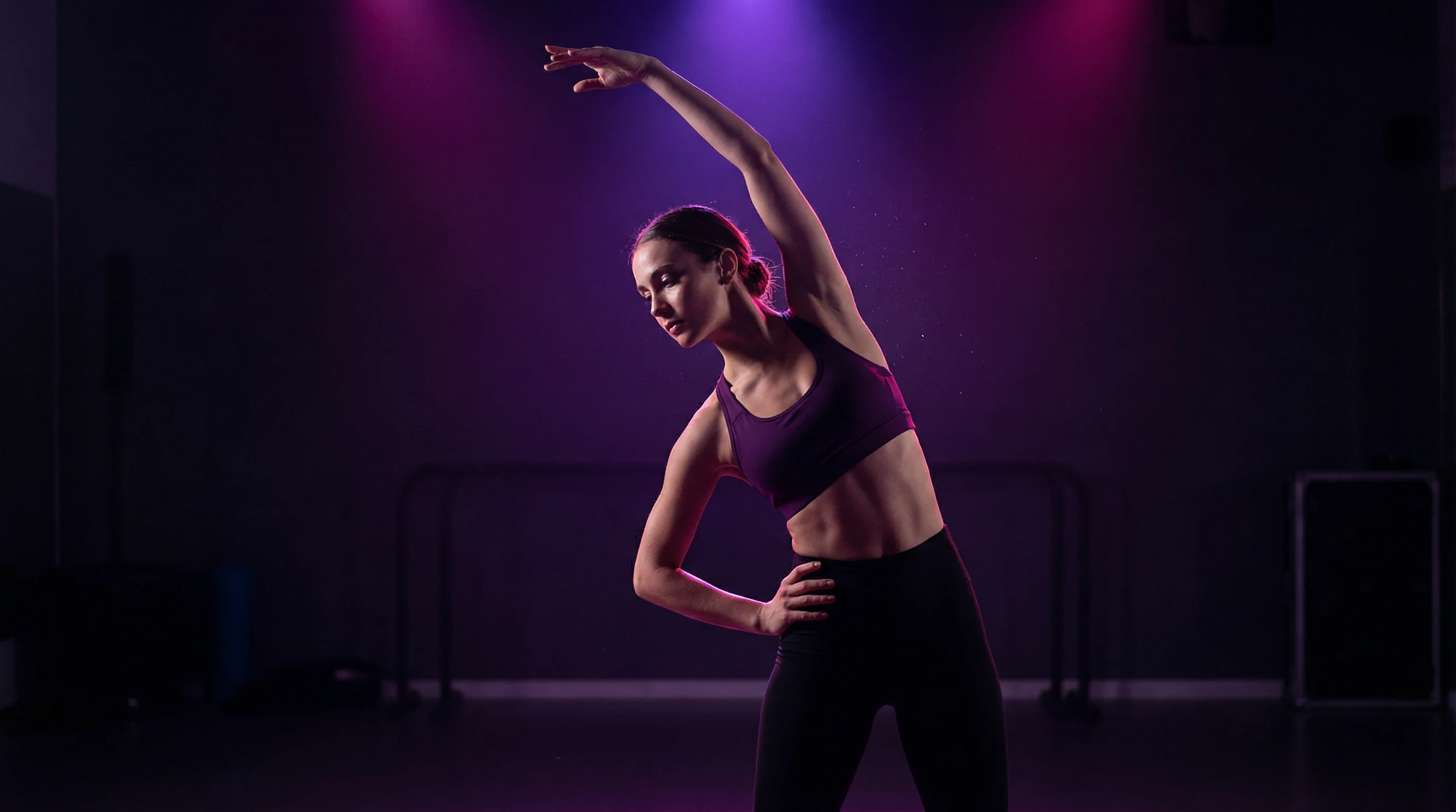 A female dancer performing a standing side stretch with one arm raised overhead in a dark studio with purple lighting