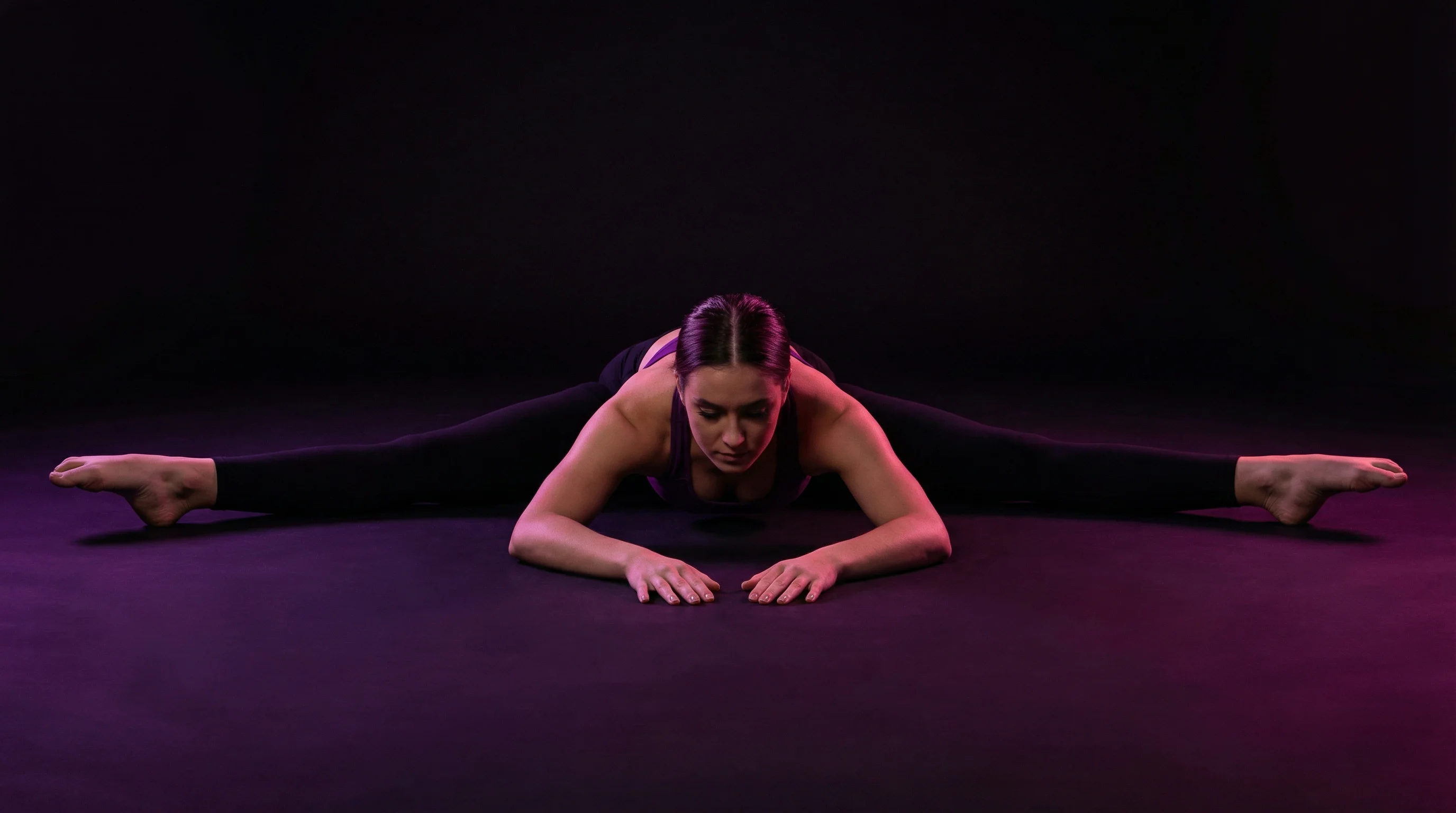 Female dancer in a perfect middle splits straddle position on a dark studio floor, leaning forward with hands on the floor