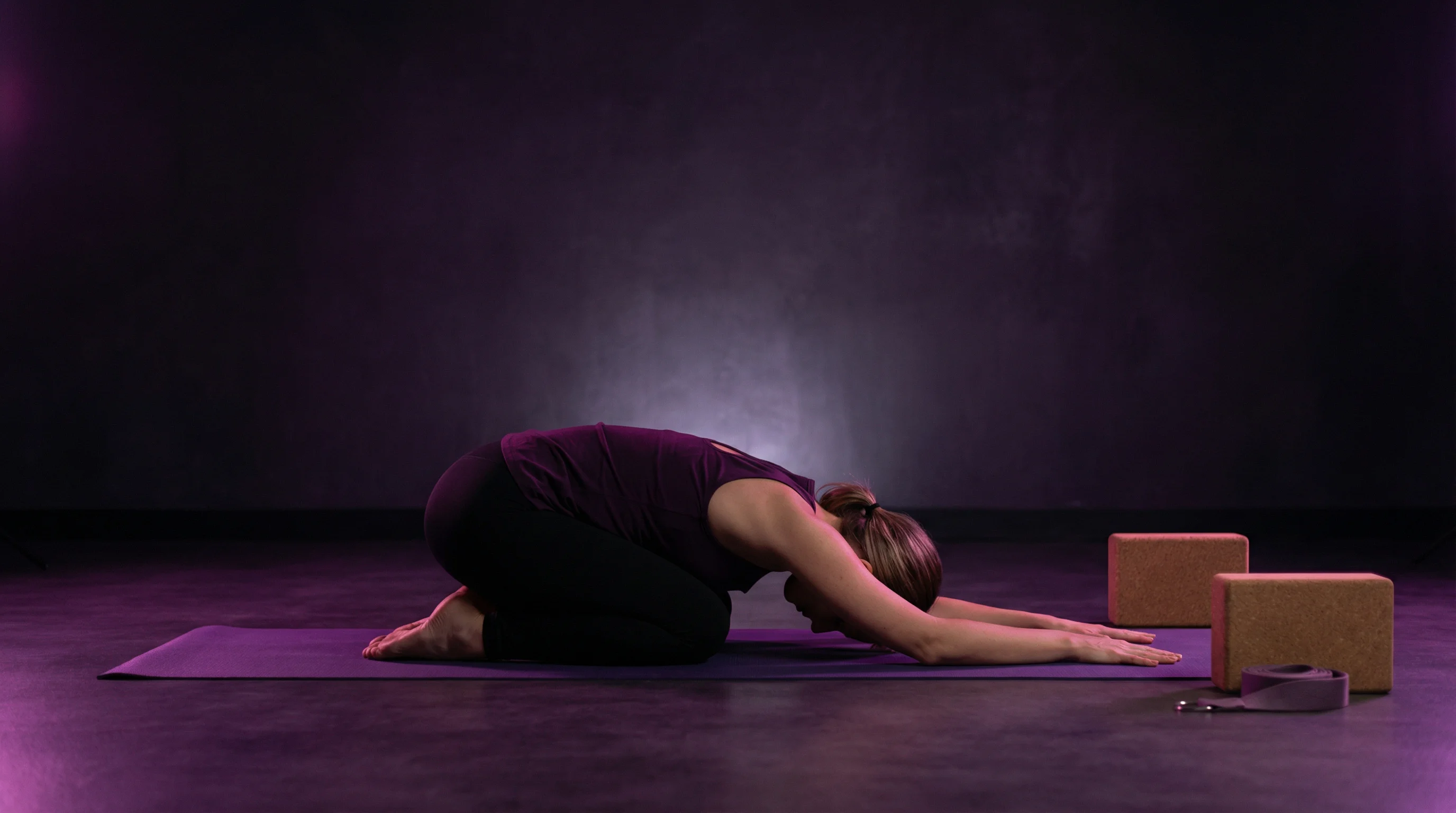 A woman in child's pose on a purple yoga mat in a dark minimalist studio with soft purple lighting